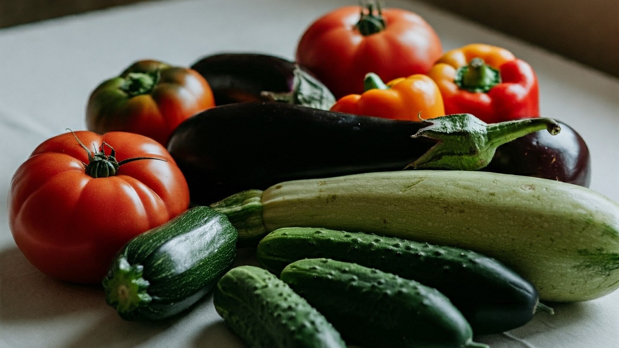 白いテーブルクロスの上に置かれた、ギリシャ料理によく使われる新鮮な野菜。トマト、キュウリ、ピーマン、ナス、ズッキーニが、それぞれの鮮やかな色合いを見せている。自然光が差し込み、野菜の瑞々しさが際立つ。